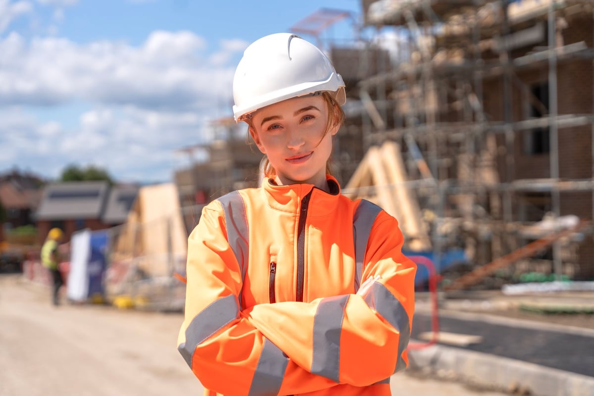 A woman in a hard hat and reflective jacket standing with crossed arms at a construction site