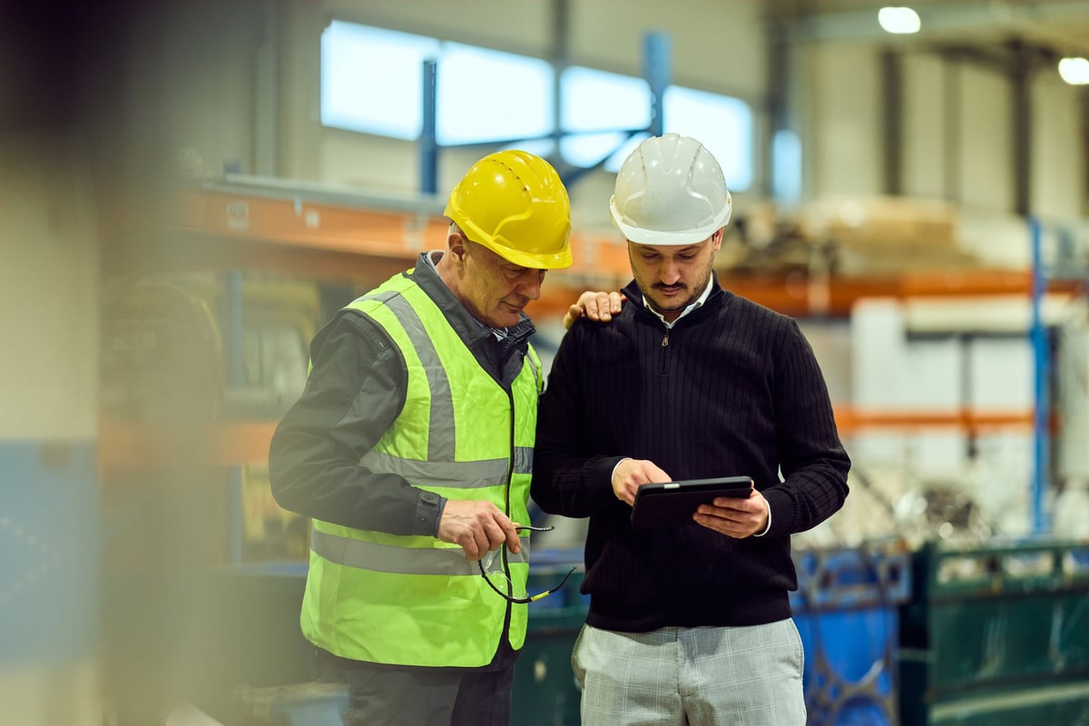 Two tradespeople in hard hats reviewing data on a tablet in a warehouse