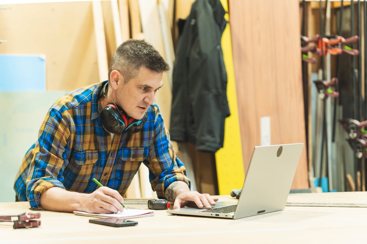 A male carpenter writing notes while working on a laptop in a woodworking shop